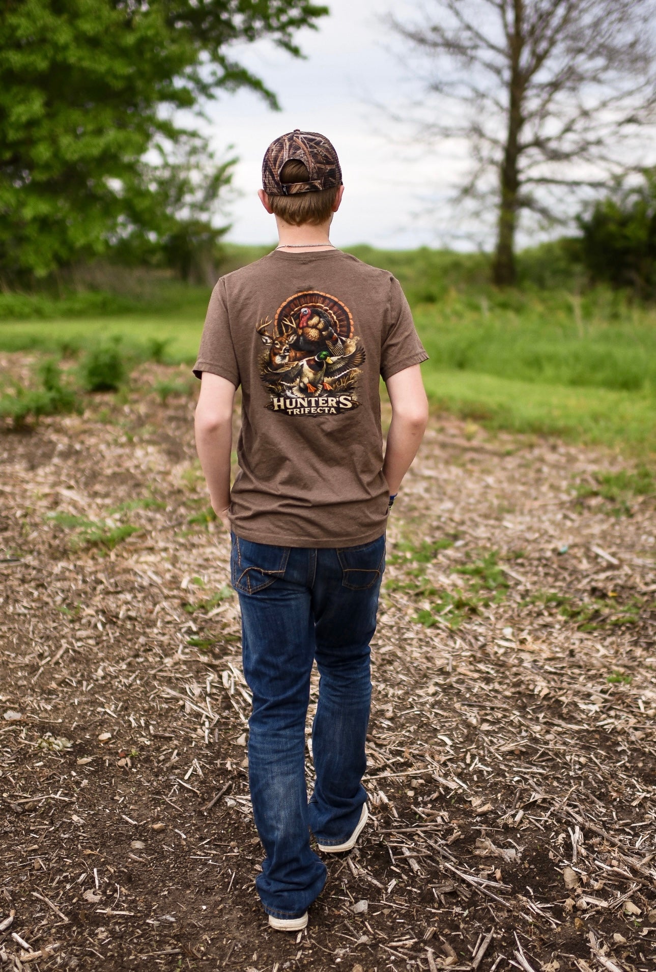 A teen wearing a brown t-shirt with a graphic design, standing in a field.